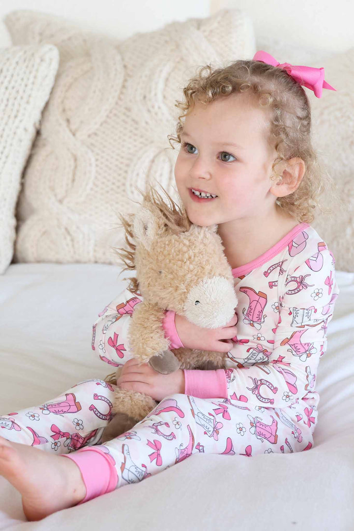 girl sitting on bed wearing a pink rodeo dreams two piece pajama set holding a stuffed horse 