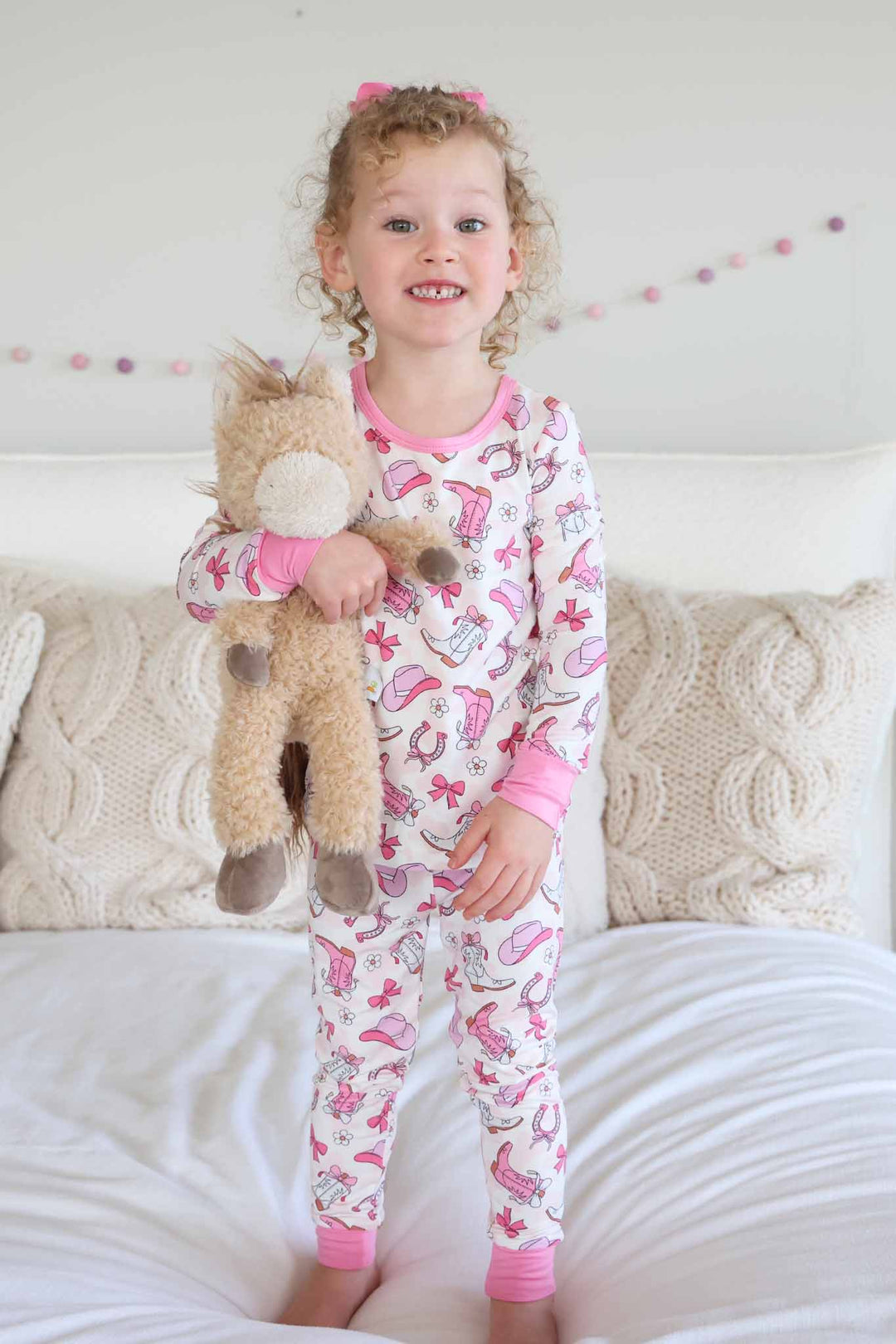 girl standing on bed in pink rodeo themed long sleeve and pant pajama set holding a stuffed horse