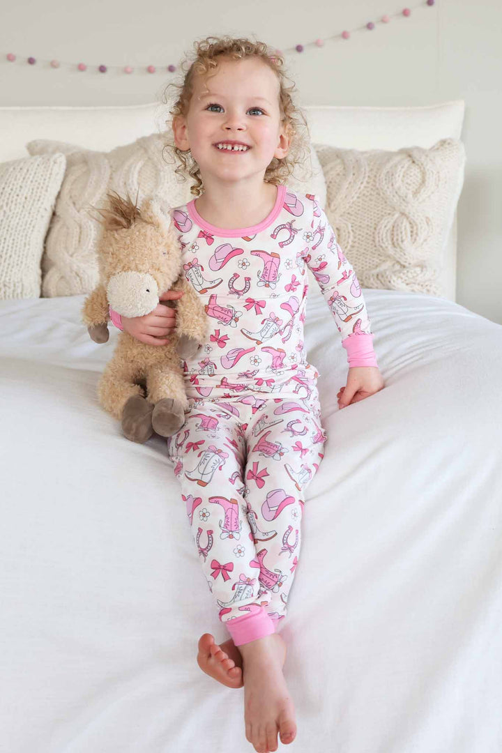 girl sitting on bed in girly western themed two piece pajama set in pink holding a stuffed horse 