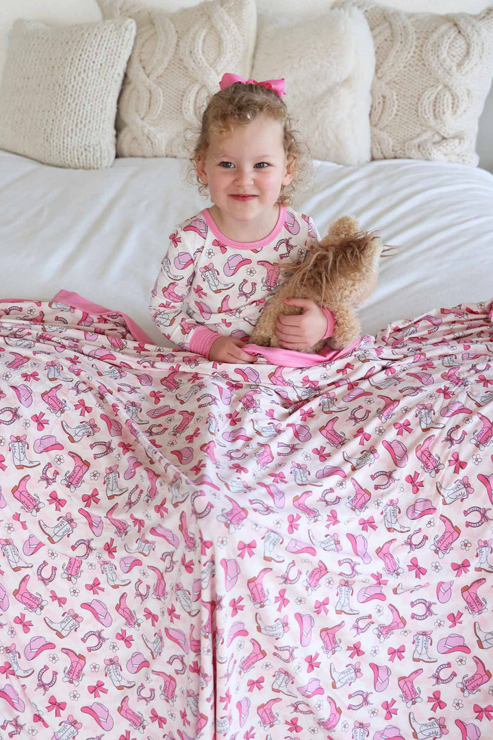 girl wearing pink rodeo dreams two piece pajama set with a matching bamboo blanket over her lap holding a horse stuffed animal 