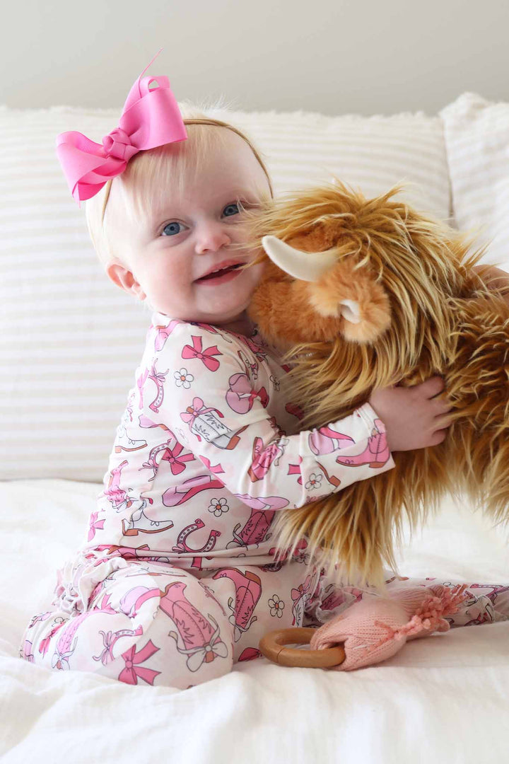 baby girl in pink western themed footie pajama hugging a stuffed highland cow
