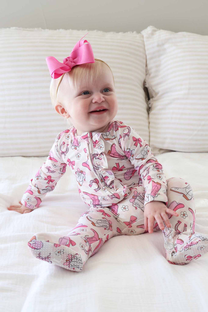 baby girl leaning back in ruffle zipper footie pajama with a girly western print and a matching hot pink ribbon bow headband 