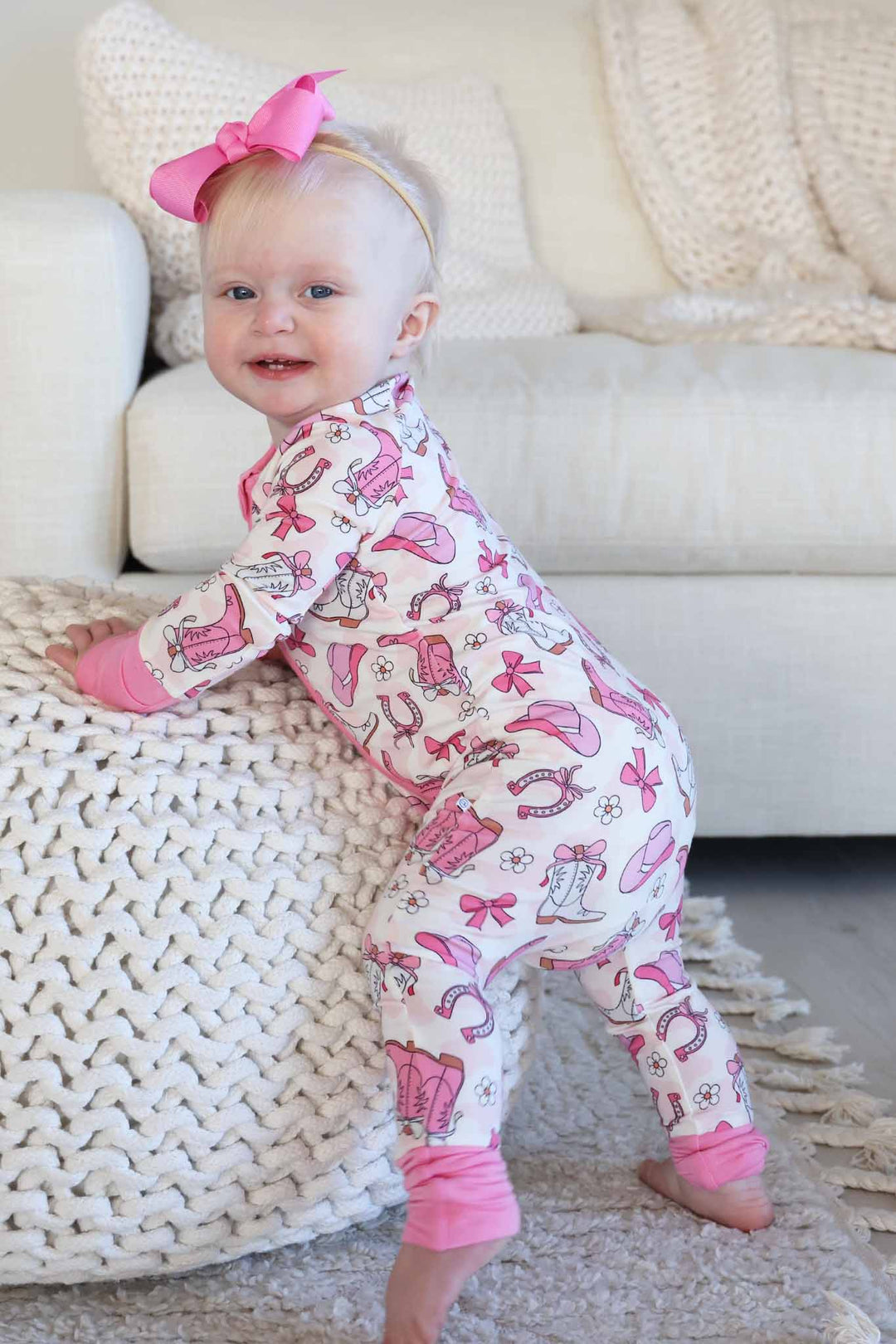 baby girl standing up wearing a girly western themed pajama romper with a matching pink ribbon bow