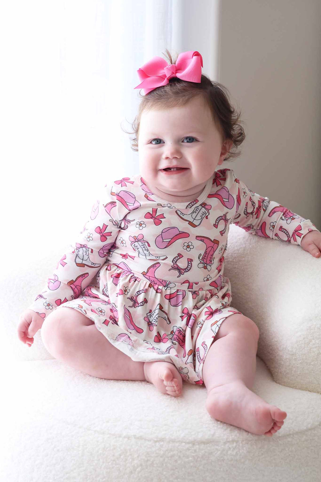 girl  sitting on white chair wearing a long sleeve skirt bodysuit with a girly cowgirl print and matching pink ribbon bow with clip