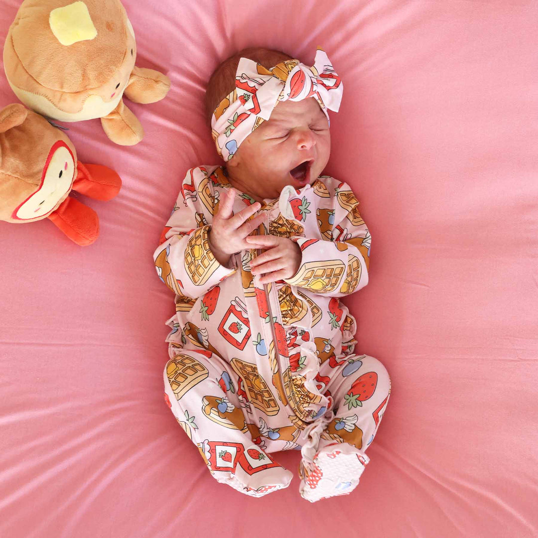 pink sweet stacks ruffle zipper footie on baby laying down wearing matching headwrap next to pancake and syrup stuffed animals 
