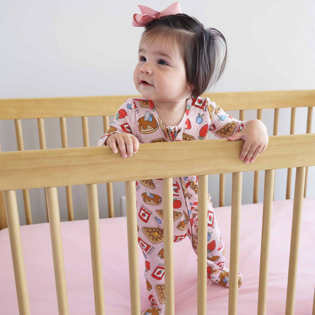 baby girl wearing pink breakfast themed footie standing in crib 