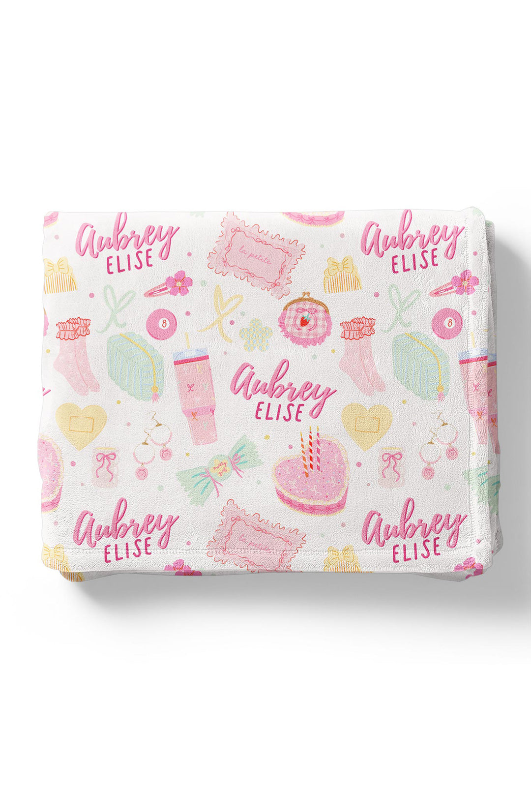 Flat lay of a folded personalized Caden Lane Pretty in Pink blanket in white, showing birthday cake, tumbler, jewelry, and custom name print