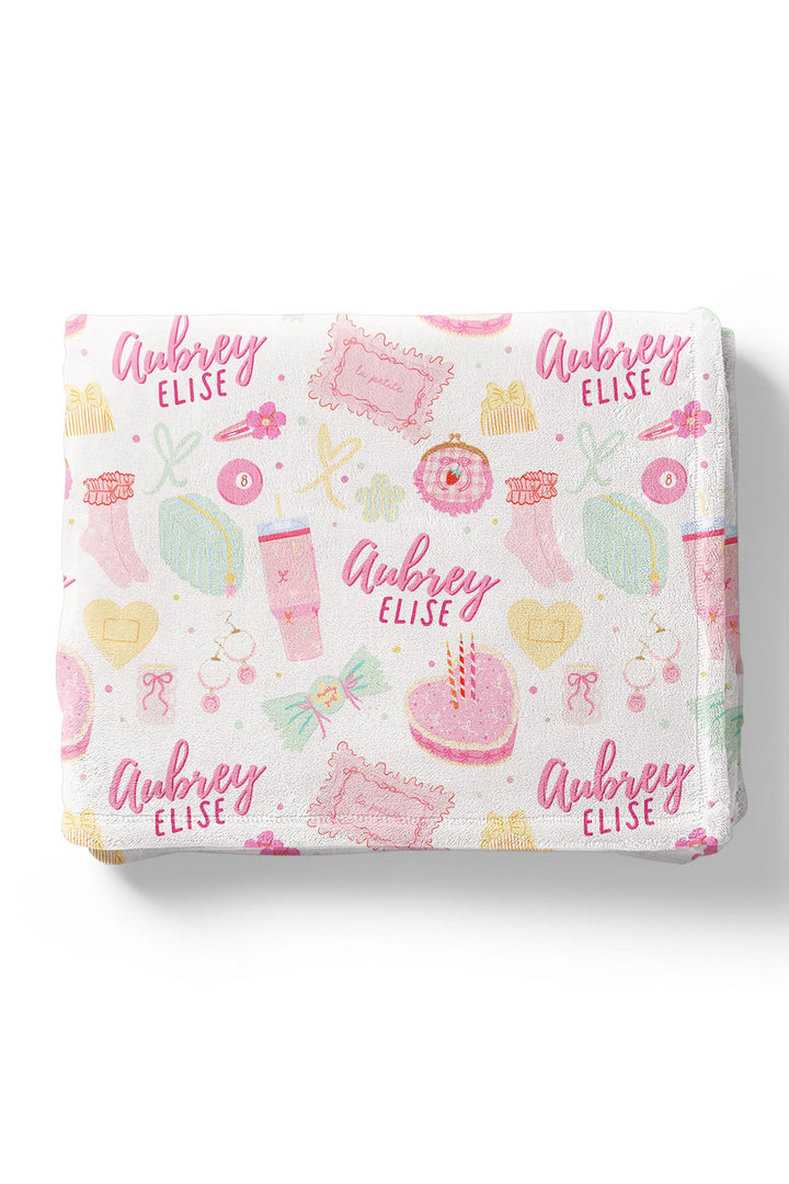 Flat lay of a folded personalized Caden Lane Pretty in Pink blanket in white, showing birthday cake, tumbler, jewelry, and custom name print