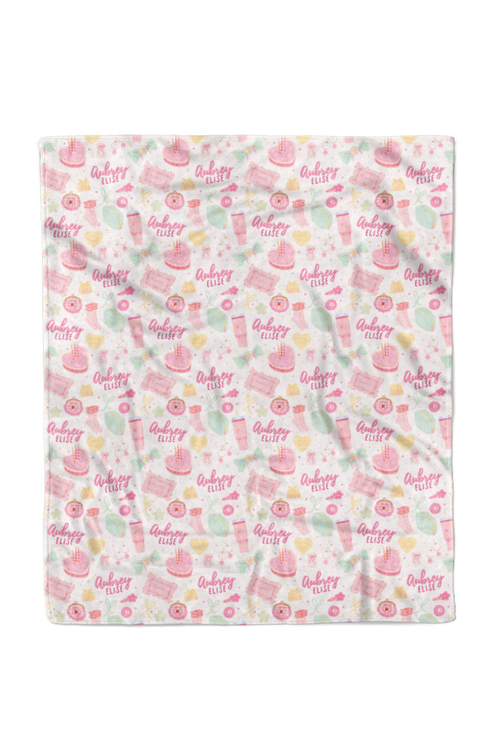 Flat lay of a fully unfolded personalized Caden Lane Pretty in Pink blanket, showing the full repeating pattern of birthday cakes, tumblers, jewelry, bows, and custom name