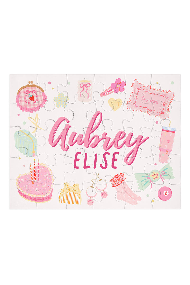 Flat lay of an assembled personalized Caden Lane Pretty in Pink jigsaw puzzle in white, showing birthday cake, jewelry, and custom name