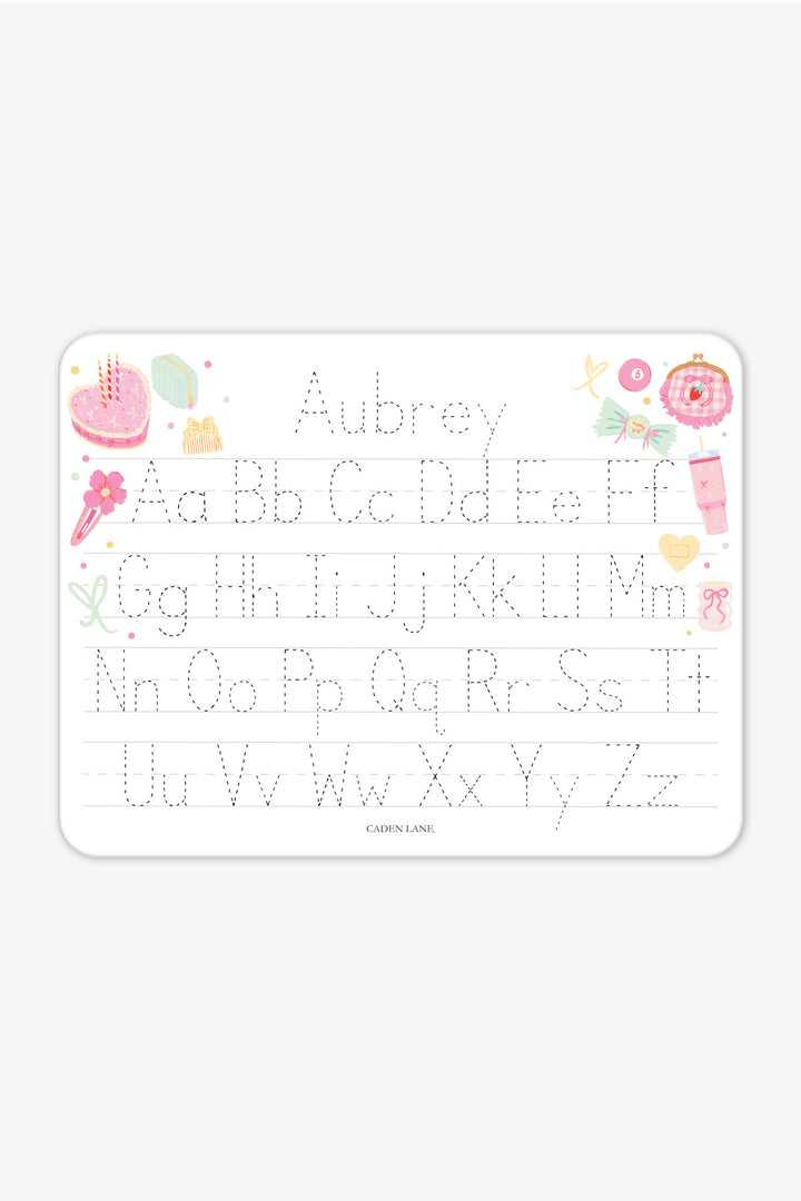 Flat lay of a personalized Caden Lane Pretty in Pink alphabet whiteboard, showing birthday cake, tumbler, and bows with tracing letters and custom name
