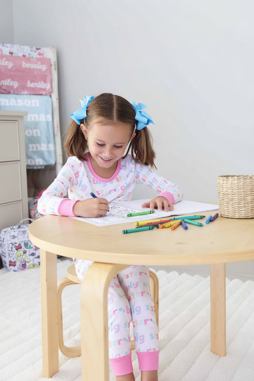 Smiling young girl with pink pigtail bows, wearing a Big Sister pajama set, coloring in a Caden Lane coloring book at a wooden table with scattered crayons and a crayon basket