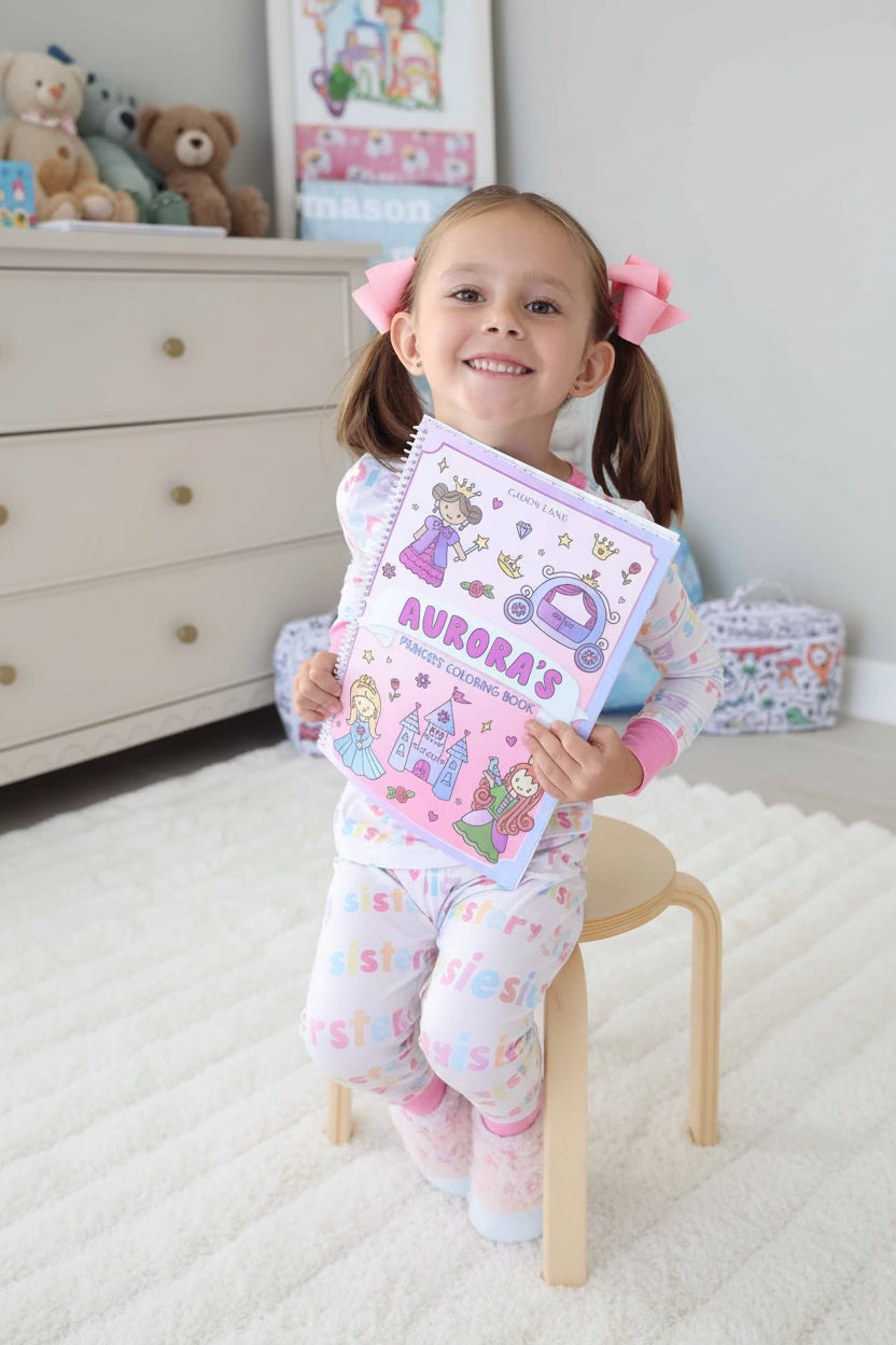 Smiling young girl with pink pigtail bows, wearing a Big Sister pajama set, holding up her personalized 'Aurora's Princess Coloring Book' by Caden Lane