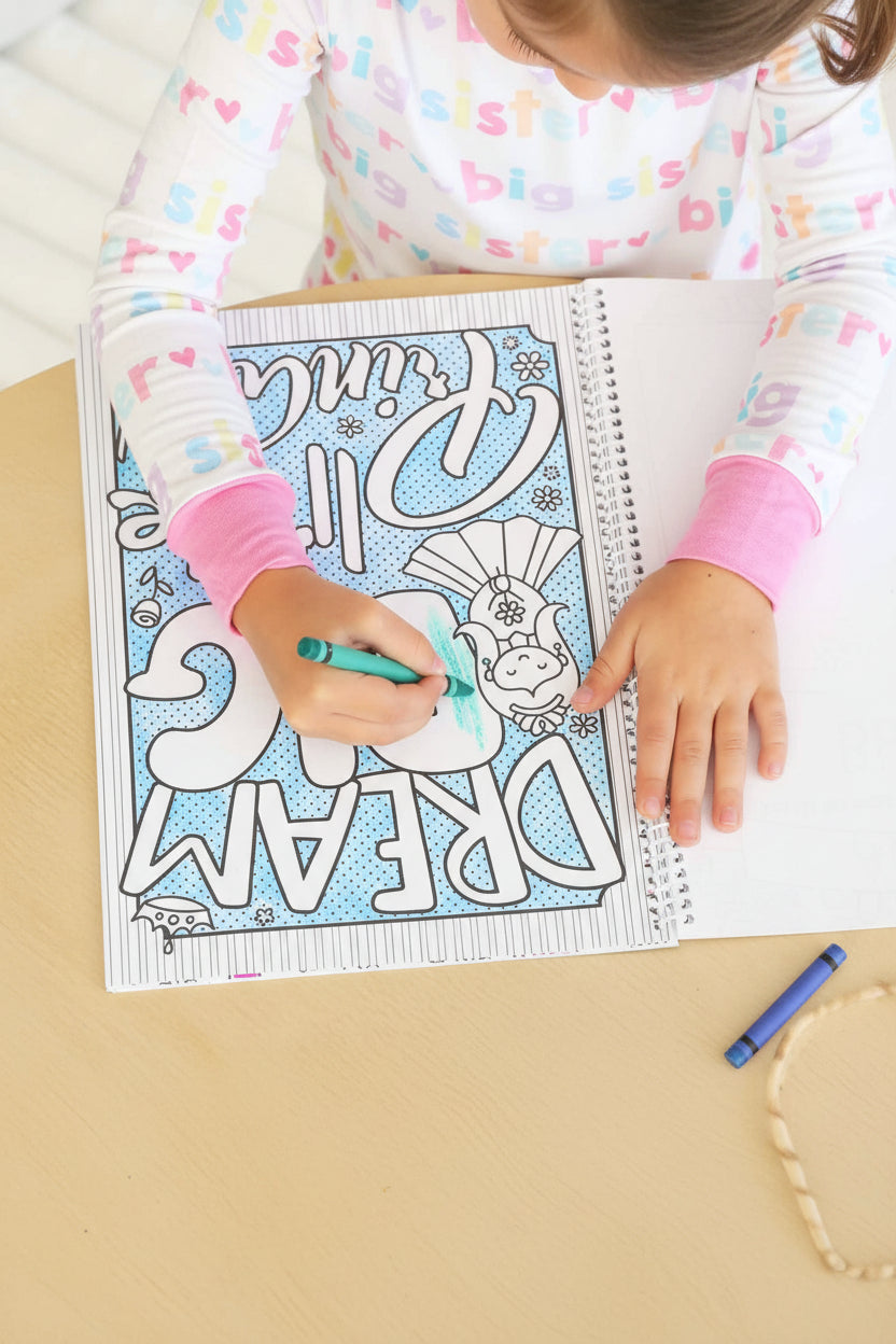 Child wearing a Big Sister pajama set coloring an upside-down 'Dream Big' page in a Caden Lane coloring book with a teal marker