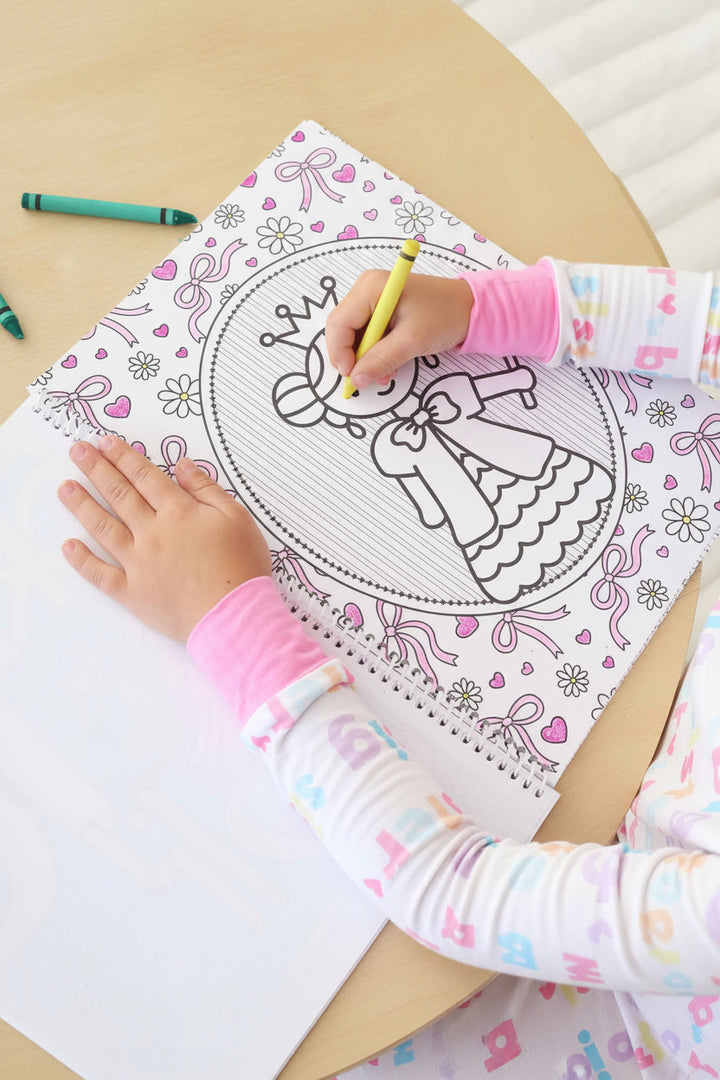 Child in a Big Sister pajama set coloring a princess page with a yellow crayon in a Caden Lane Princess Coloring Book, framed by a bow and floral border