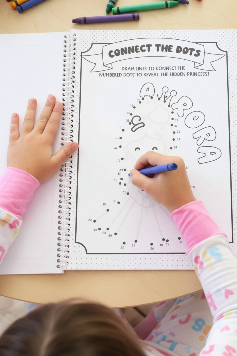 Child in a Big Sister pajama set connecting numbered dots with a blue crayon to reveal a hidden princess on a personalized 'Aurora' activity page.