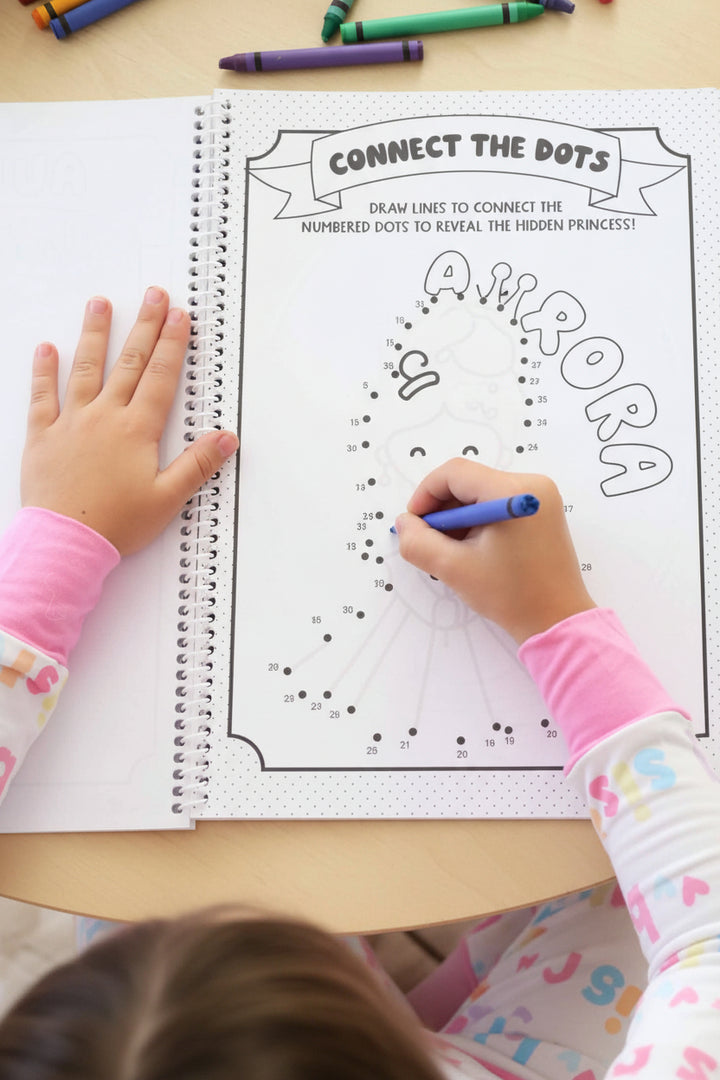 Child in a Big Sister pajama set connecting numbered dots with a blue crayon to reveal a hidden princess on a personalized 'Aurora' activity page.