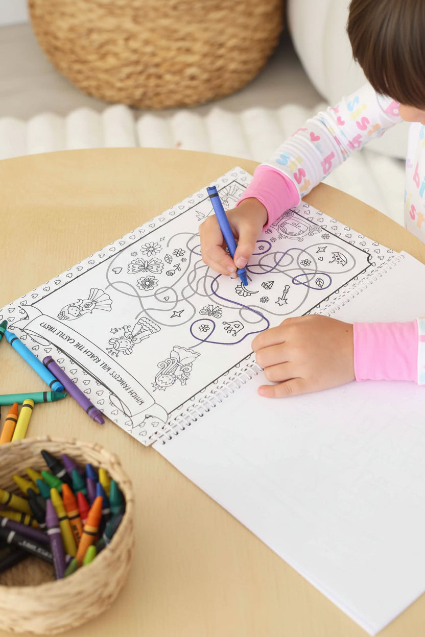 Child in a Big Sister pajama set tracing a maze path with a blue crayon on a 'Which princess will reach the castle first?' activity page in a Caden Lane coloring book
