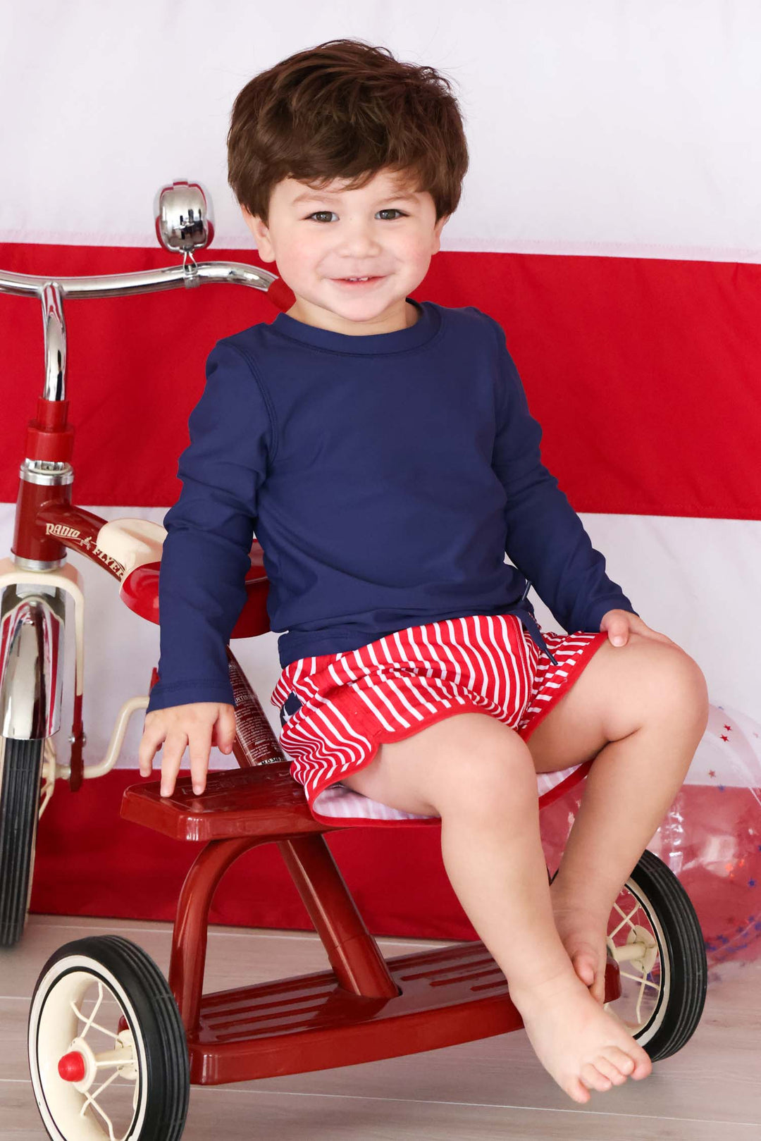 boy sitting on tricycle in navy rash guard shirt and red and white striped trunks
