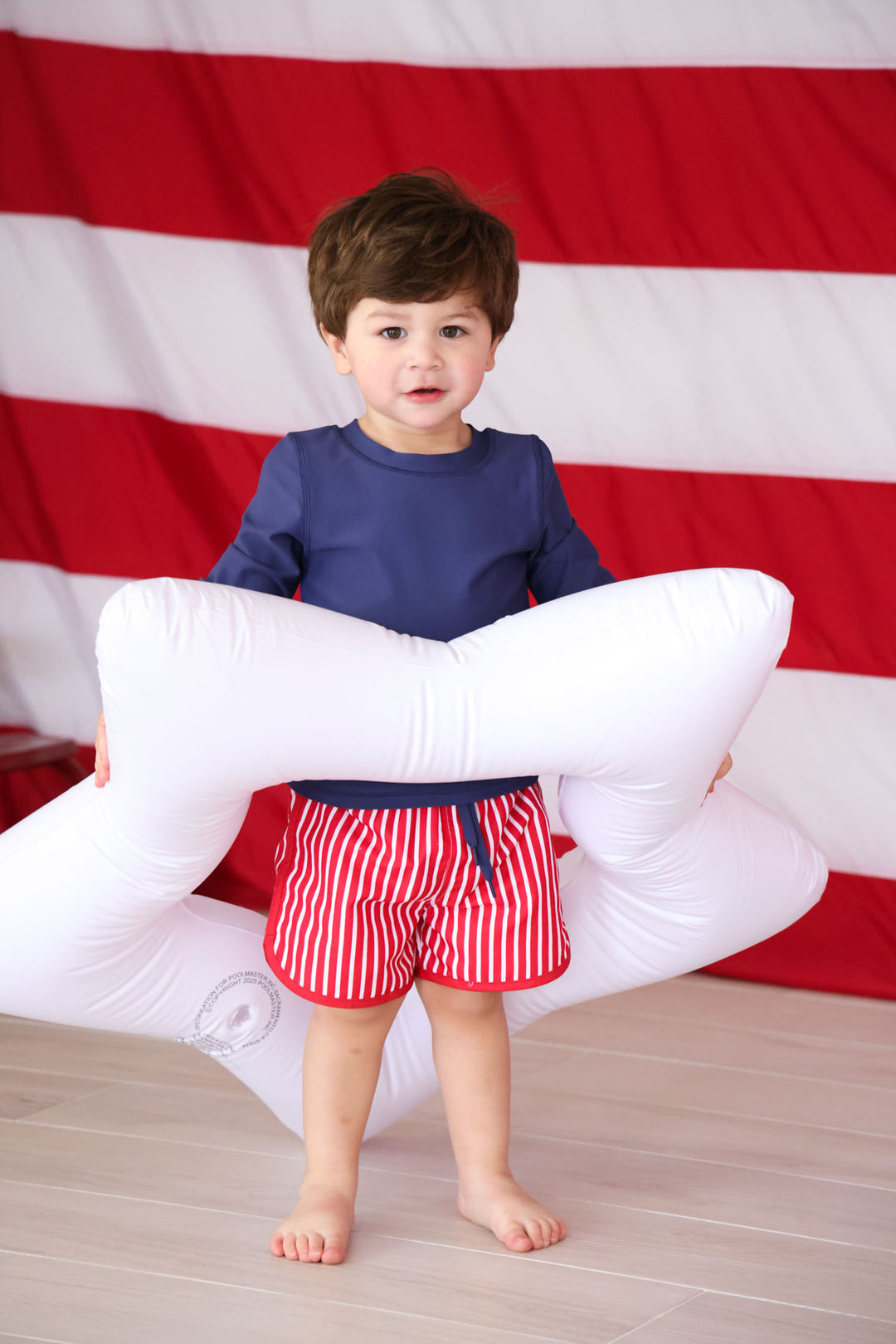 boy standing in white star innertube with a navy long sleeve shirt and red and white swim trunks 