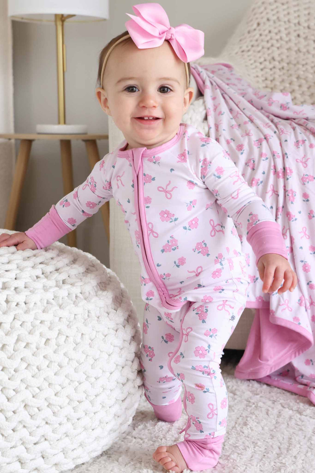 smiling standing baby girl in pink ribbon and roses bamboo viscose convertible zip romper with matching light pink ribbon bow headband 