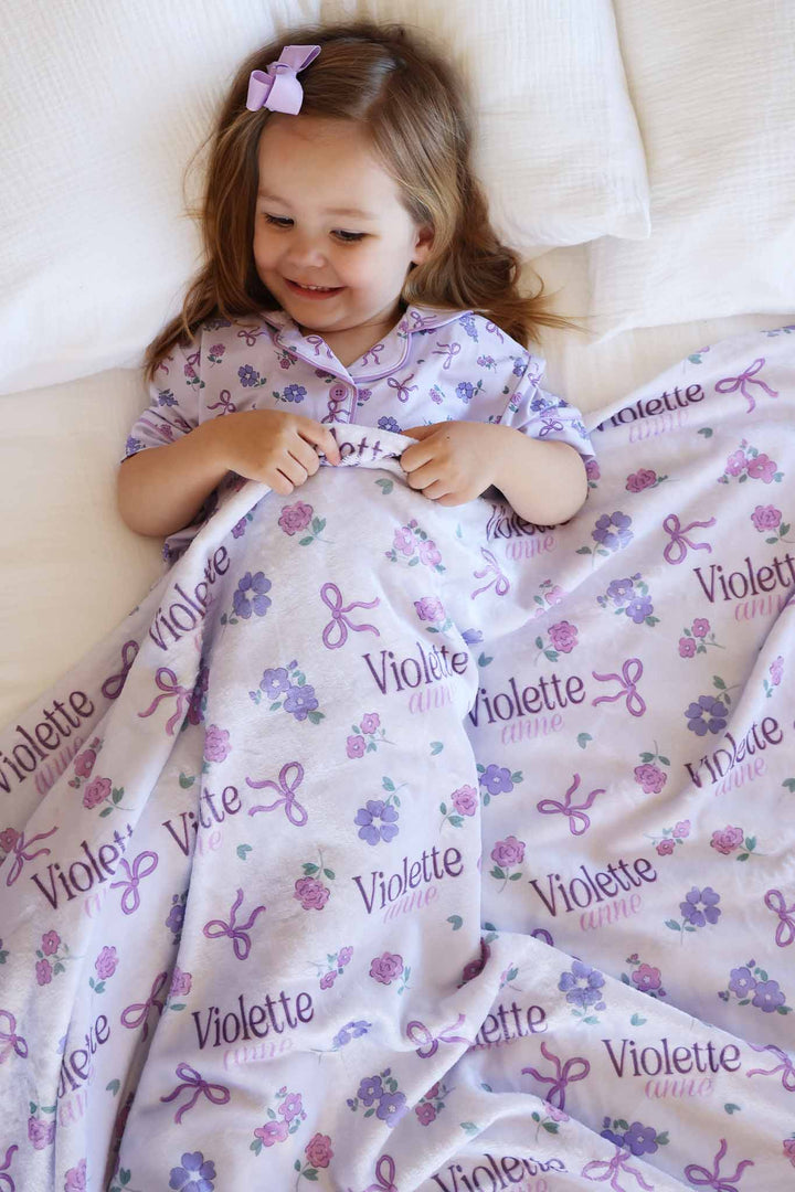 girl laying on bed with personalized flower and ribbon purple personalized blanket over her 