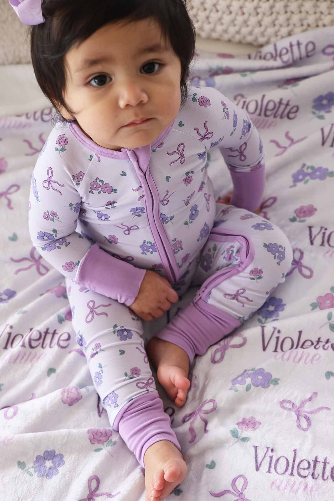 baby girl sitting on bed wearing purple ribbon and roses convertible zip romper on top of matching personalized name blanket 