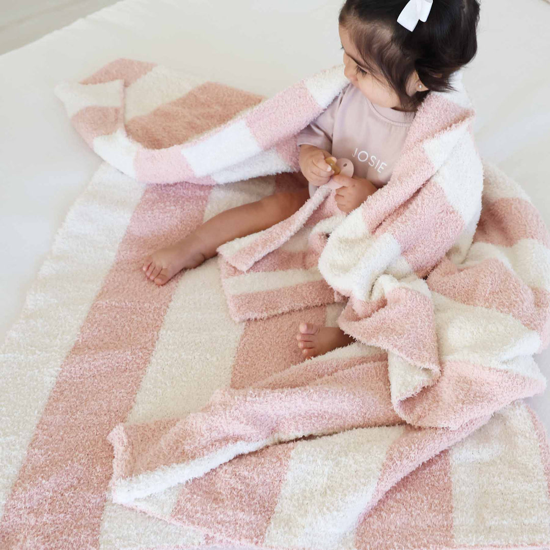 girl sitting on a bed with a pink and white stripe microfiber blanket draped over her holding a matching lovey 