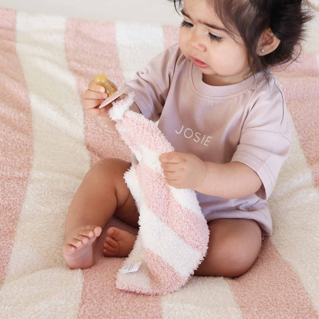 girl sitting on a bed wearing a personalized tshirt romper for babies and toddlers holding a pacifier attached to a pink and white stripe plush lovey and sitting on a matching blanket 