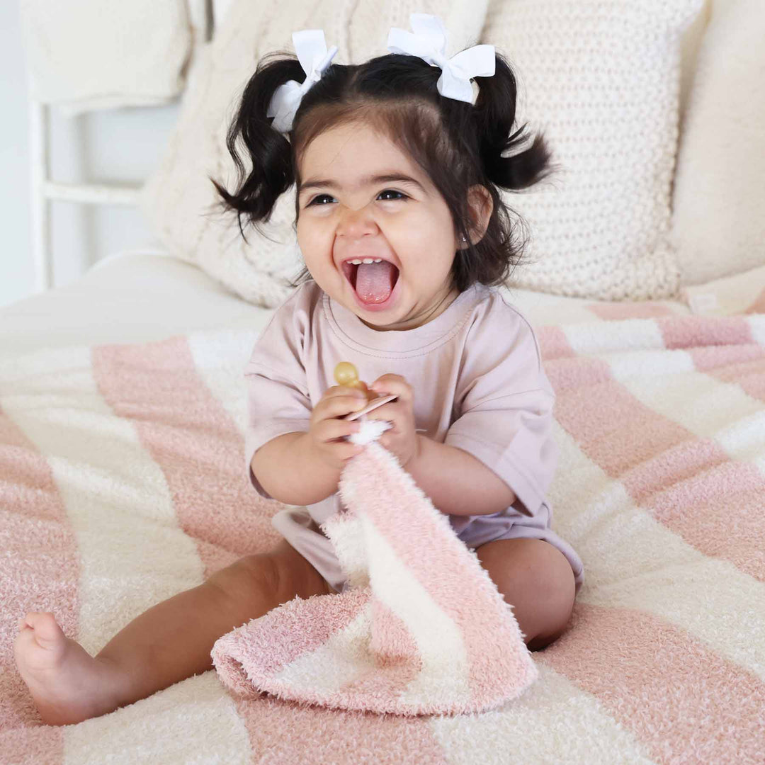 girl sitting on a bed holding a pink and white stripe plush lovey with a pacifier 