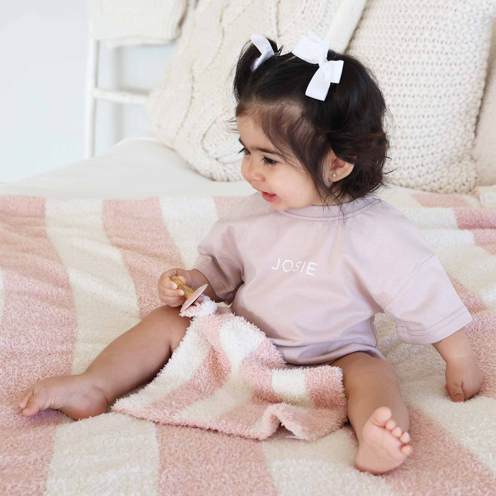 baby girl sitting on a bed wearing a pink and white stripe microfiber blanket holding a matching pink and white stripe lovey