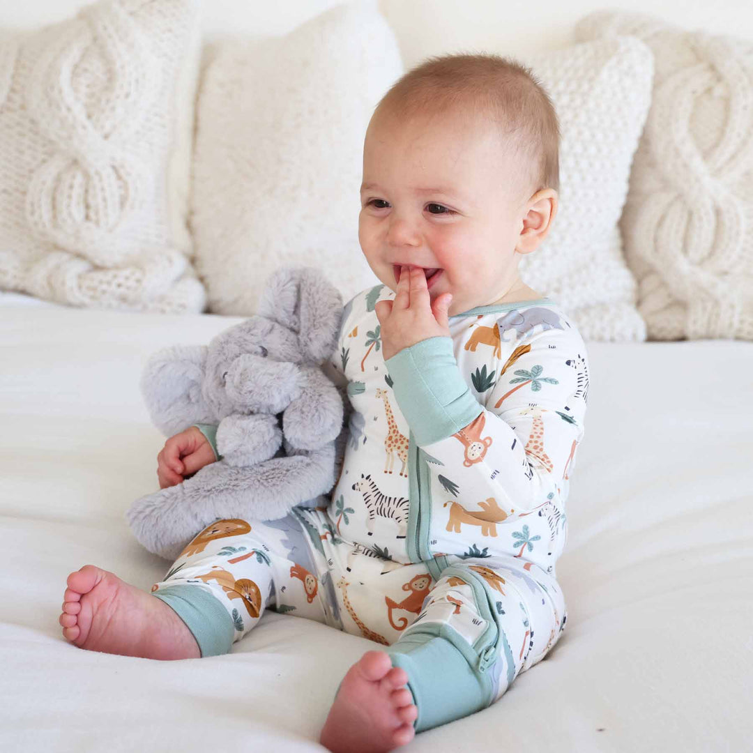 baby sitting on bed in safari pajama romper holding a grey plush elephant lovey