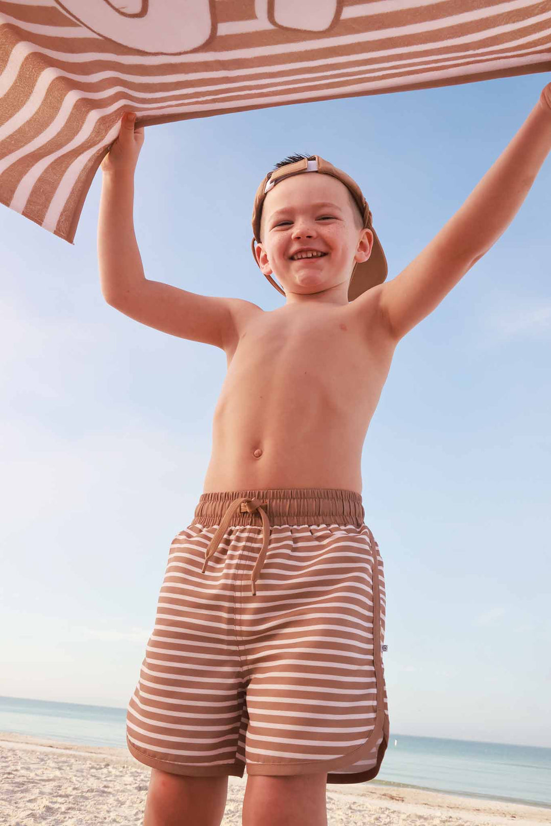 boy standing on sand wearing brown and white stripes swim trunks with a matching waterproof baseball cap waving a towel in the air 