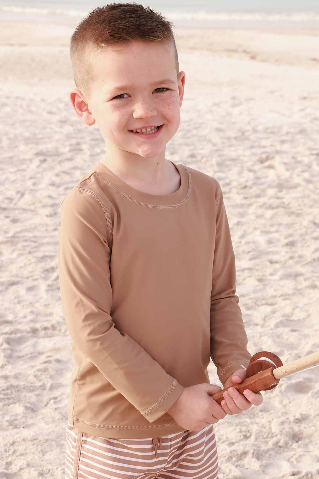 boy standing on the beach in solid brown rash guard shirt with matching striped swim trunks holding a fishing pole 