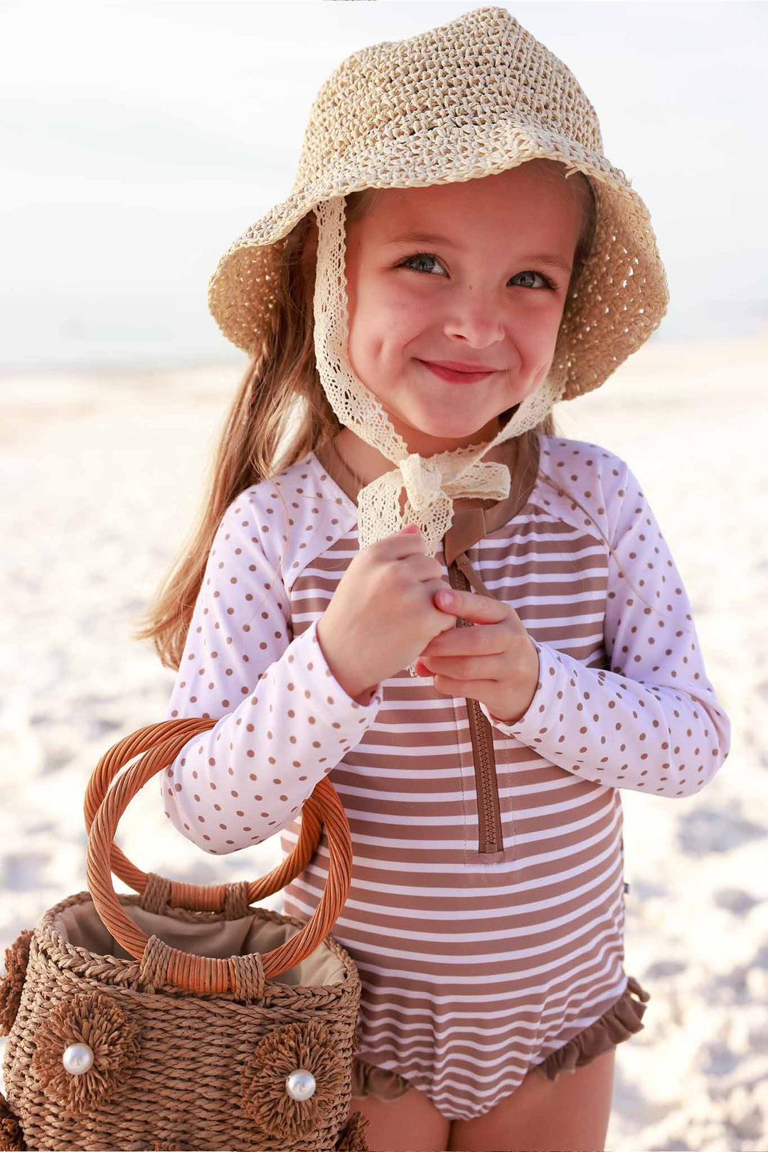girl standing on the beach in brown and white stripe and polka dot rash guard swimsuit holding a rattan purse and sun hat 