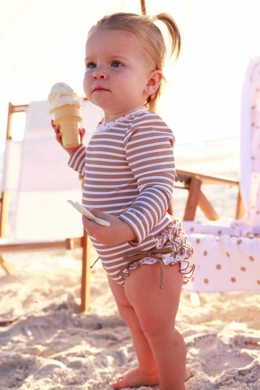 baby girl standing on the beach in brown and white stripe long sleeve ruffle bottom rash guard romper holding a vanilla ice cream cone