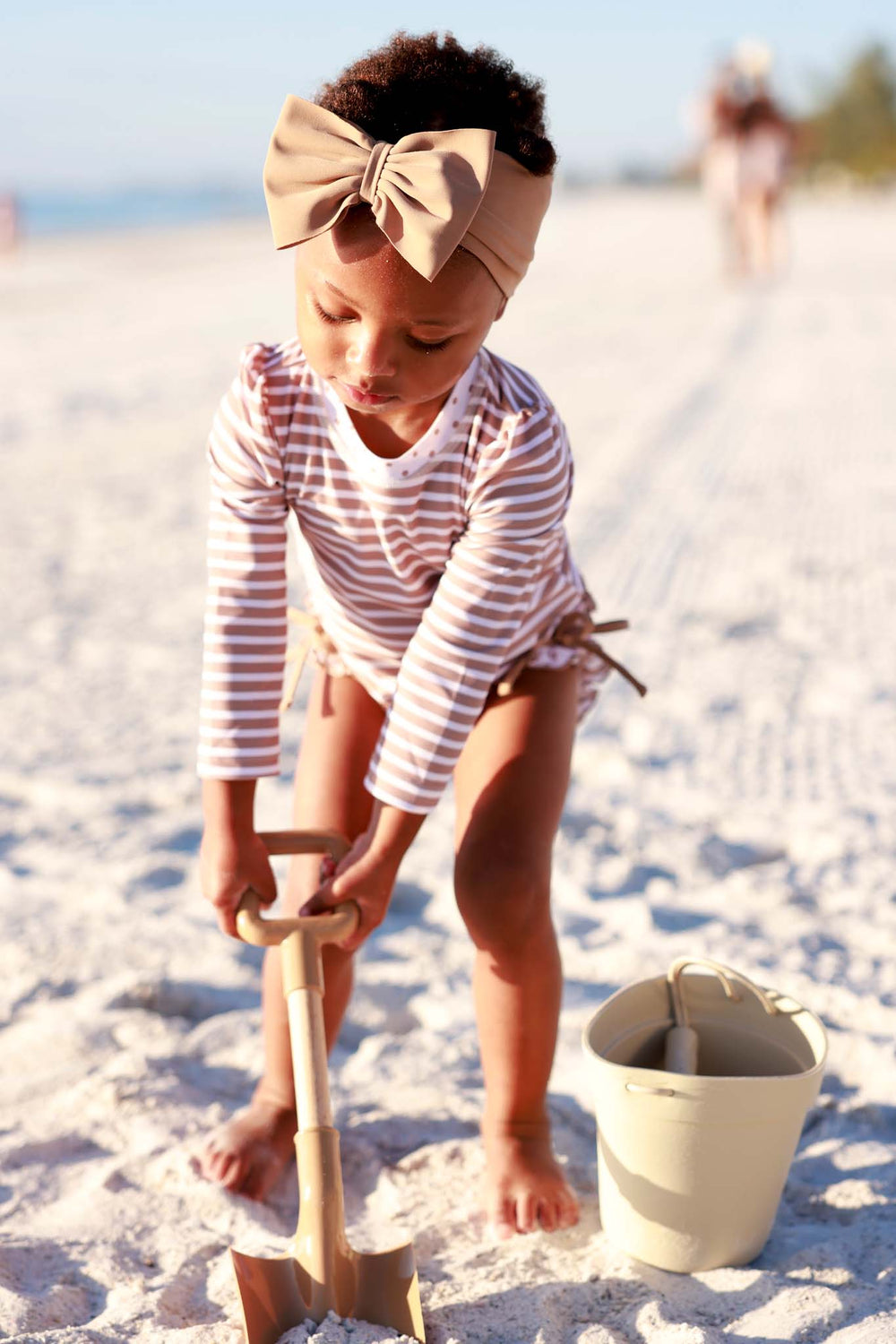 girl digging a hold on the beach wearing a brown and white stripe rash guard swimsuit with a matching brown swim bow 