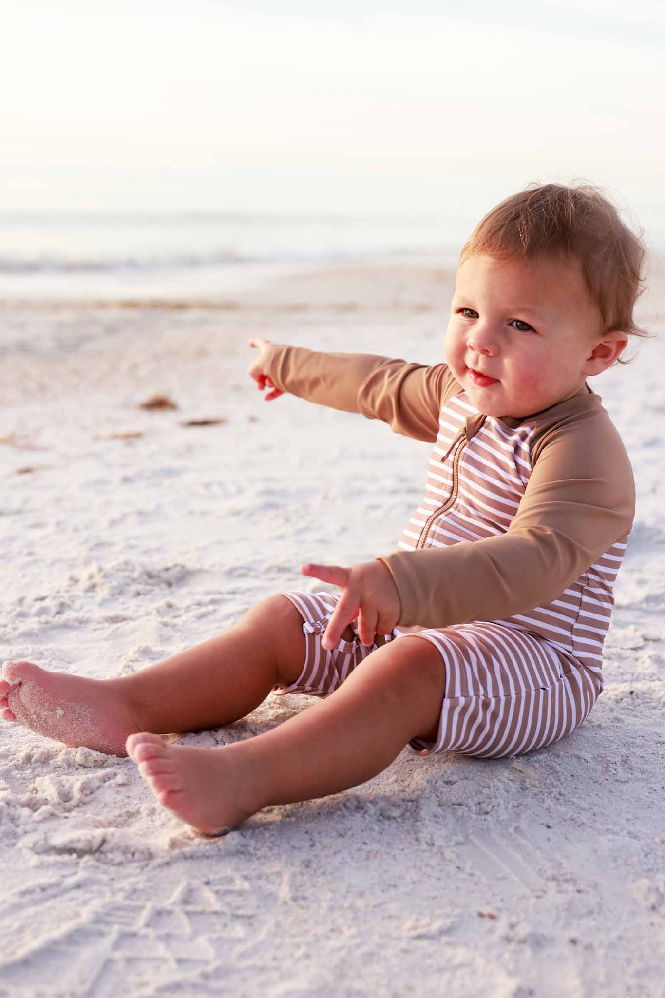 boy sitting on beach wearing a brown and white stripe swim romper shortie with solid brown long sleeves and a front zipper 