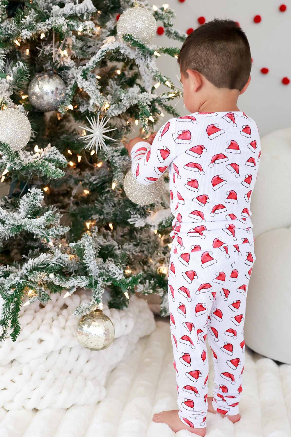 Boy in a Caden Lane two-piece Christmas pajama set with a festive Santa hat print decorating a beautifully lit Christmas tree