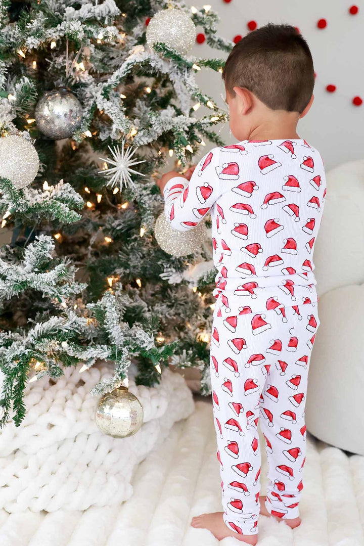 Boy in a Caden Lane two-piece Christmas pajama set with a festive Santa hat print decorating a beautifully lit Christmas tree