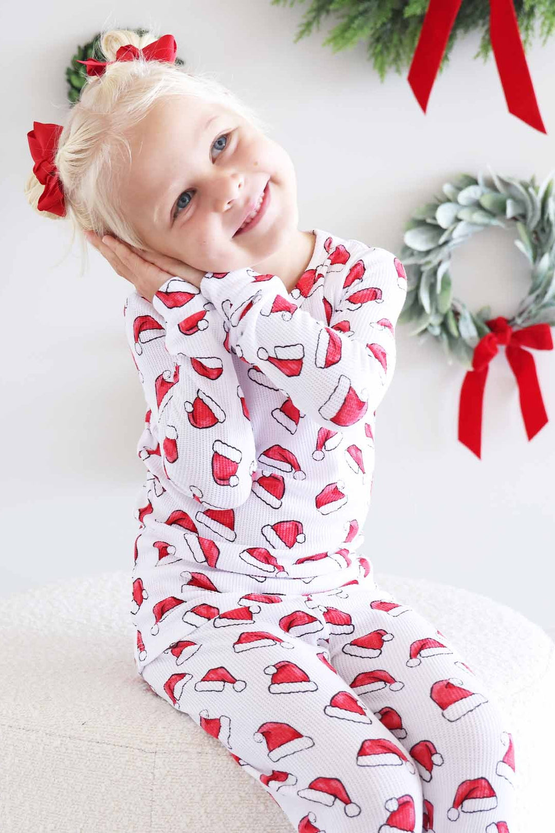 Girl wearing Caden Lane white two-piece holiday pajamas with a festive red Santa hat print poses with hands tucked under their cheek, surrounded by Christmas wreaths and red bows for a cozy holiday portrait