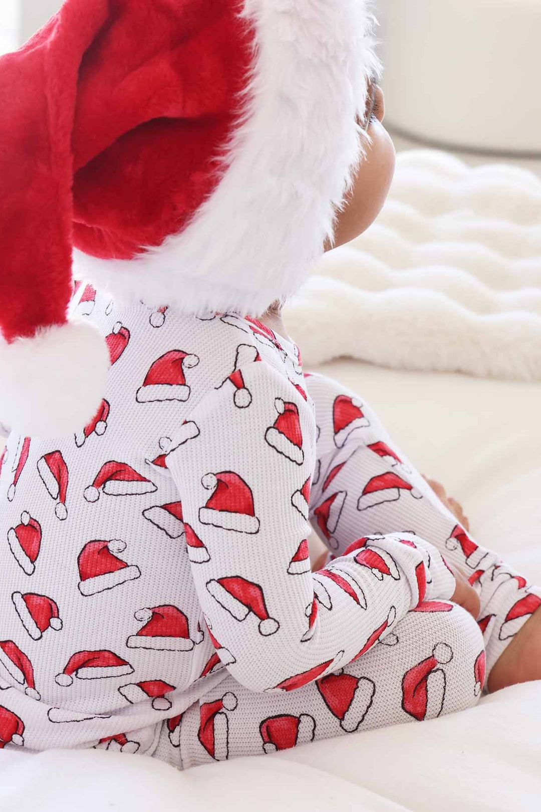 Child wearing Caden Lane white two-piece holiday pajamas with a red Santa hat print sits on a cozy white blanket while wearing a plush Santa hat, creating a festive Christmas morning look