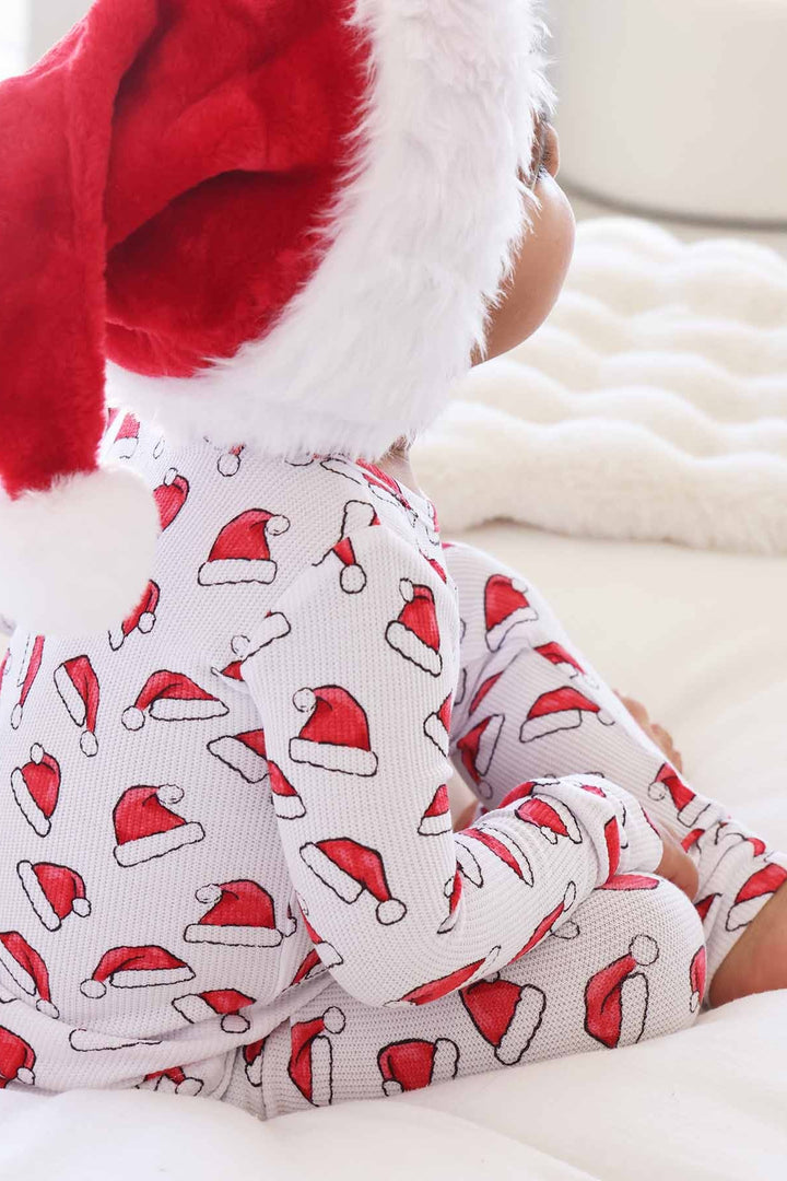 Child wearing Caden Lane white two-piece holiday pajamas with a red Santa hat print sits on a cozy white blanket while wearing a plush Santa hat, creating a festive Christmas morning look
