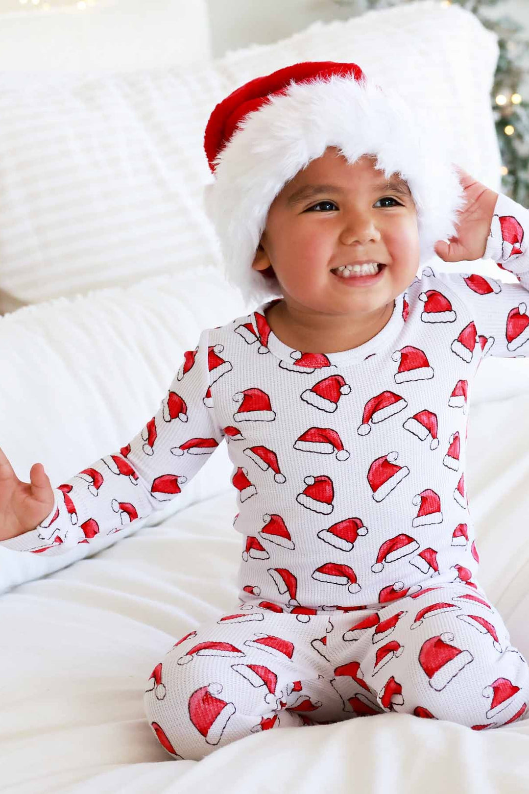 Child wearing Caden Lane white two-piece holiday pajamas with a red Santa hat print sits on a cozy white blanket while wearing a plush Santa hat, creating a festive Christmas morning look