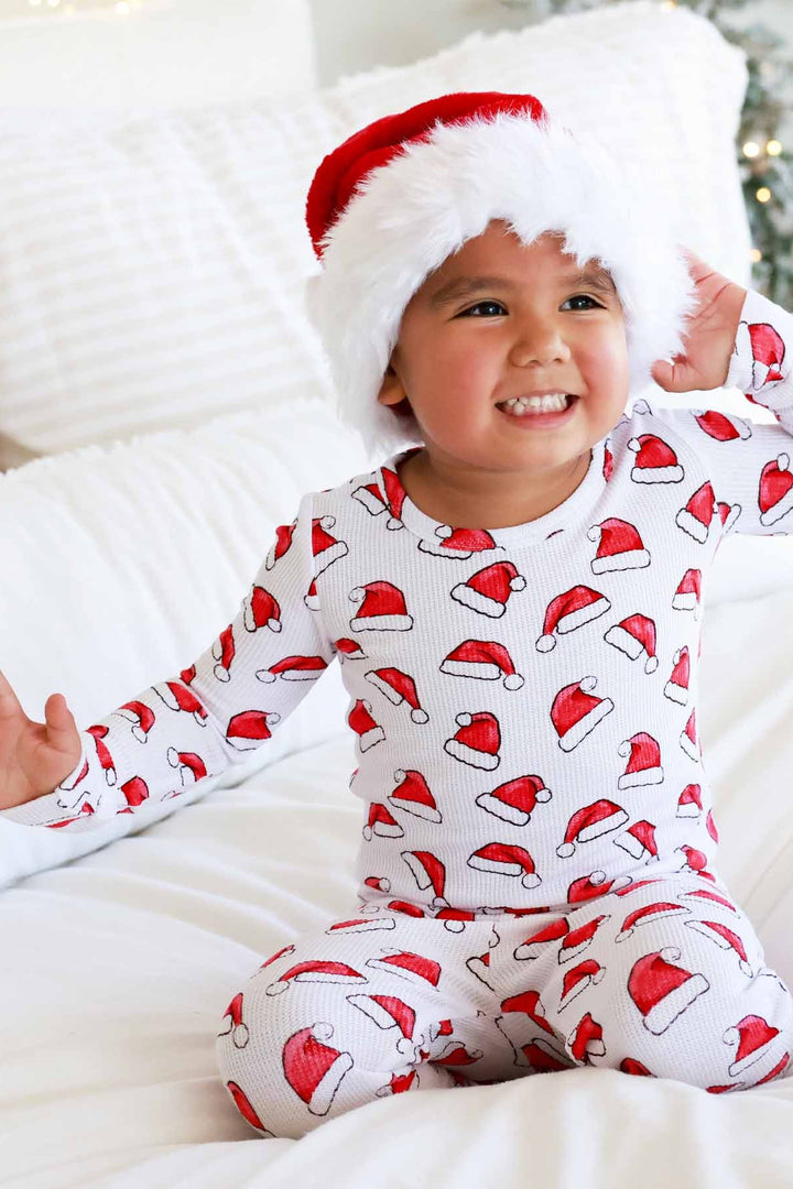 Child wearing Caden Lane white two-piece holiday pajamas with a red Santa hat print sits on a cozy white blanket while wearing a plush Santa hat, creating a festive Christmas morning look