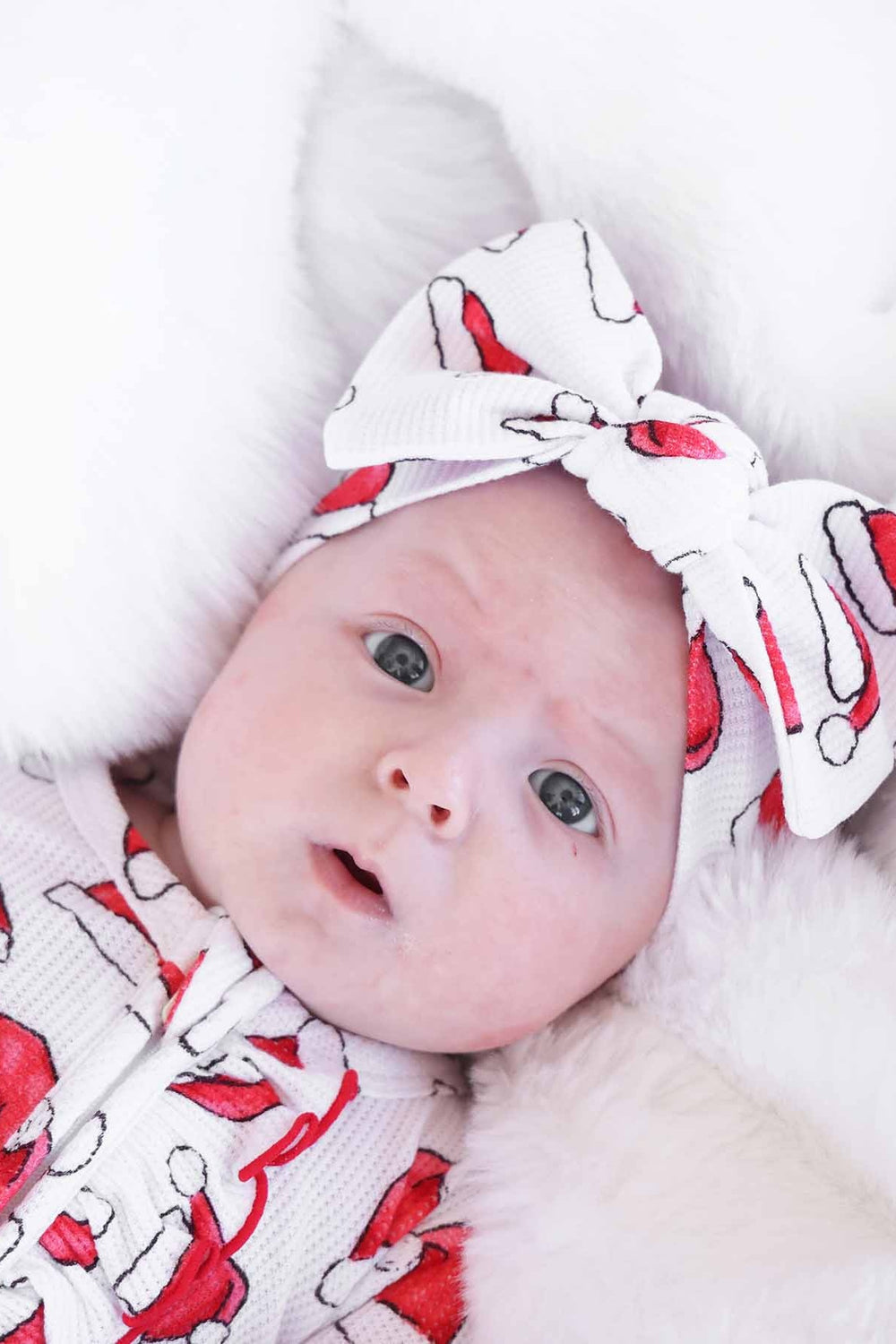 Close-up of a baby wearing a Caden Lane white waffle-knit zip romper and matching knotted headwrap with a festive red Santa hat print, lying on a soft white faux fur blanket for a cozy holiday look