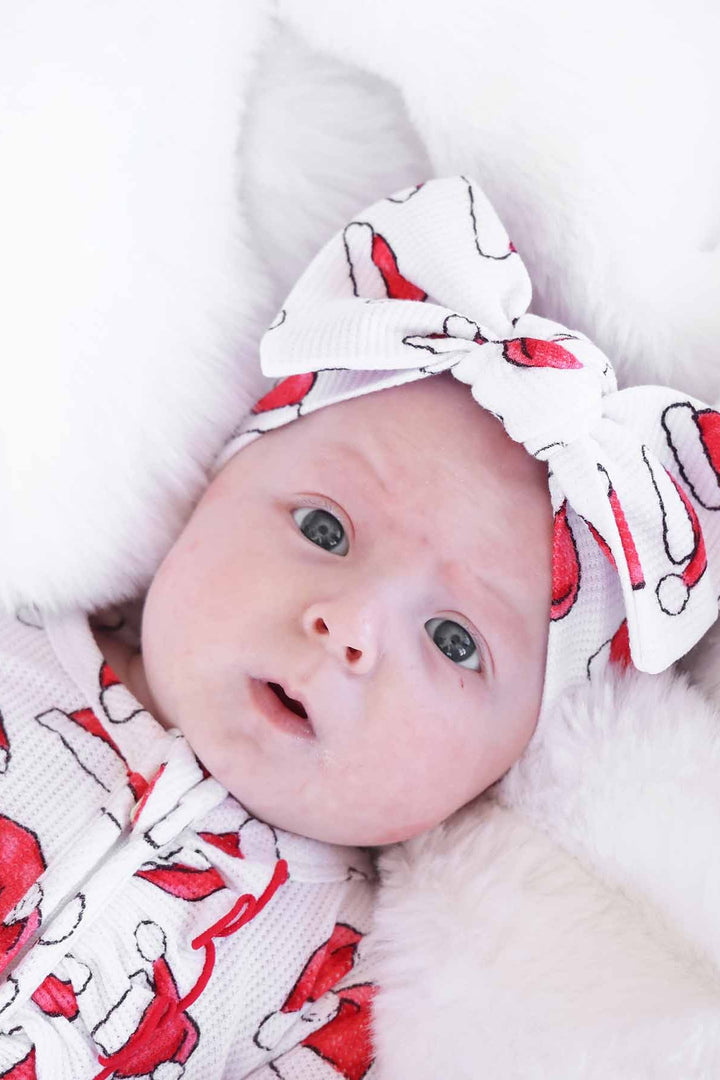 Close-up of a baby wearing a Caden Lane white waffle-knit zip romper and matching knotted headwrap with a festive red Santa hat print, lying on a soft white faux fur blanket for a cozy holiday look