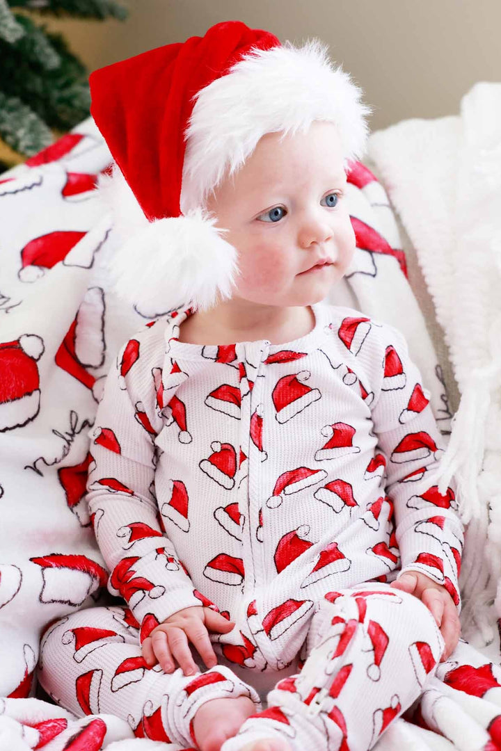 Toddler wearing a Caden Lane white waffle-knit zip romper with a festive red Santa hat print sits on a cozy blanket wearing a plush Santa hat, creating a classic Christmas morning look