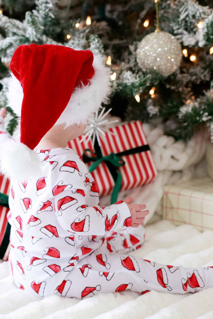 Toddler wearing a Caden Lane white waffle-knit zip romper with a festive red Santa hat print sits on a cozy blanket wearing a plush Santa hat