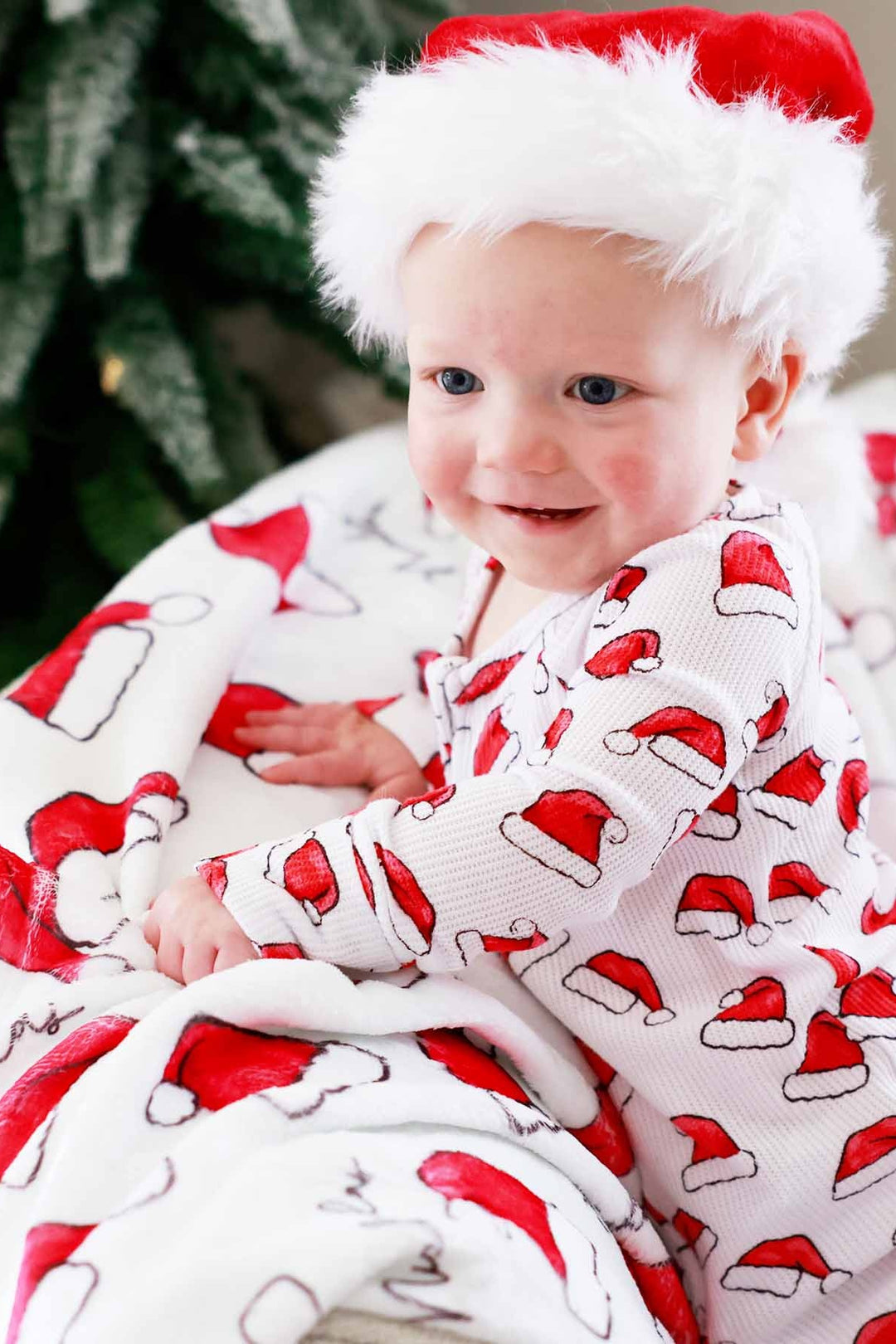 Toddler wearing a Caden Lane white waffle-knit zip romper with a festive red Santa hat print sits on a cozy blanket wearing a plush Santa hat